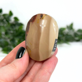 Hand holding a brown mookaite palm stone with a blurred green and white background