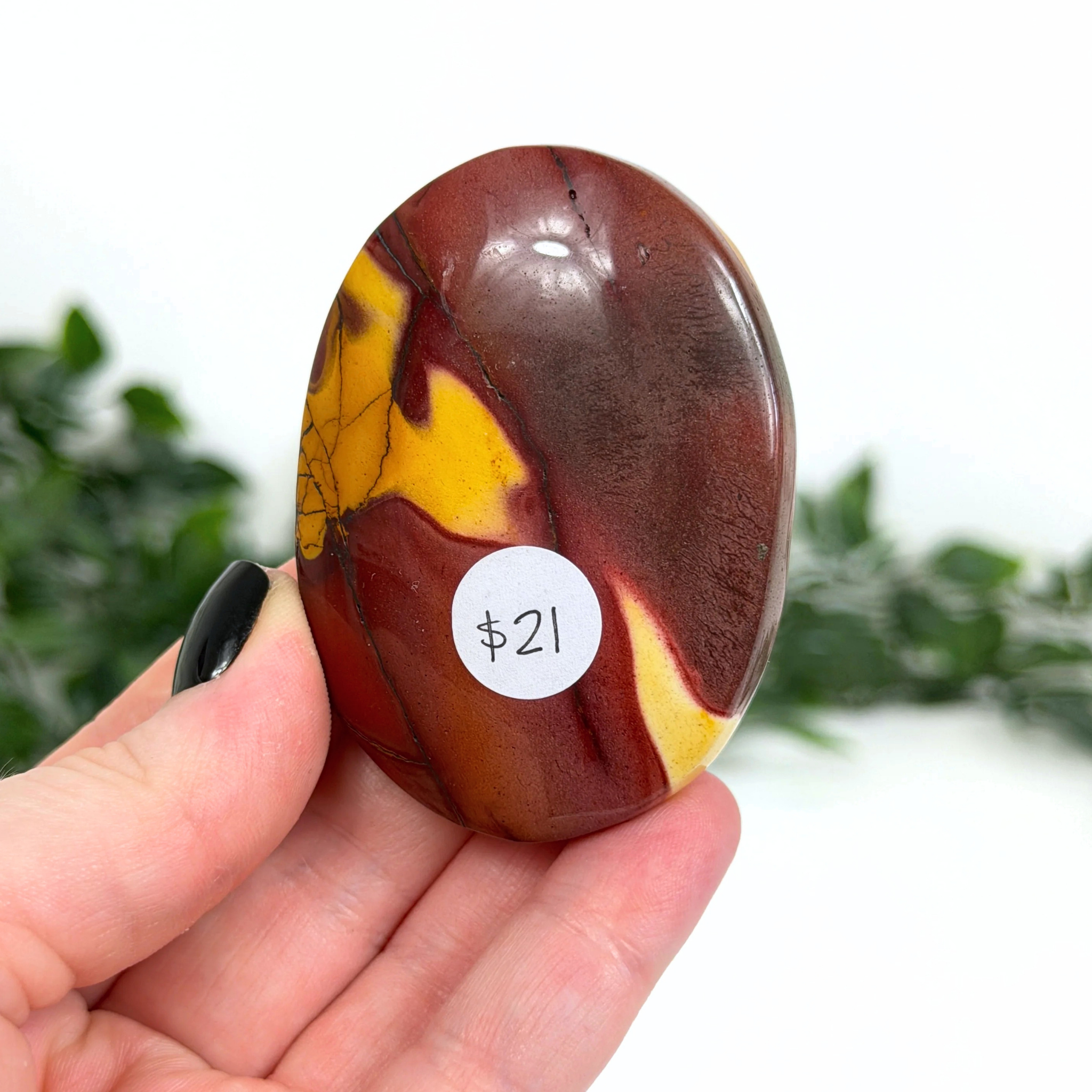 Red and yellow mookaite palm stone held in a hand with a price tag of $21, against a blurred green background