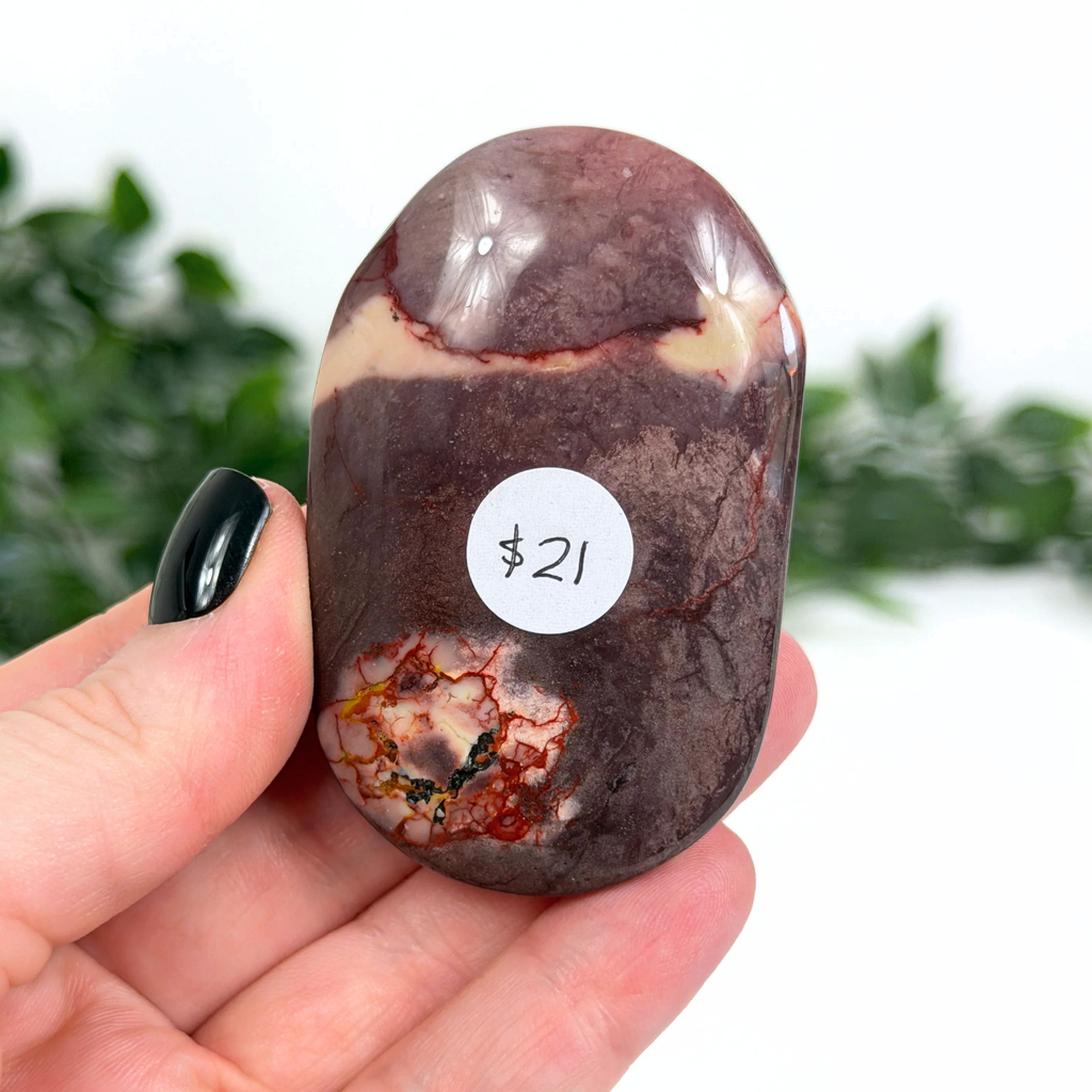 Purple mookaite palm stone held in a hand with a price tag of $21, against a blurred green and white background.
