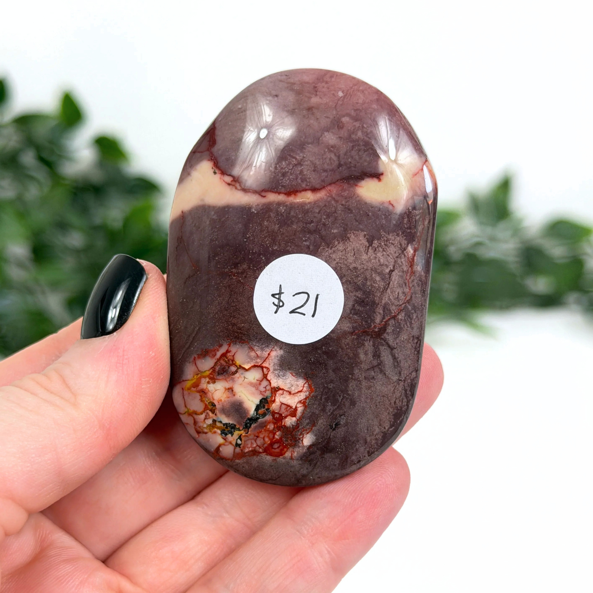 Purple mookaite palm stone held in a hand with a price tag of $21, against a blurred green and white background.