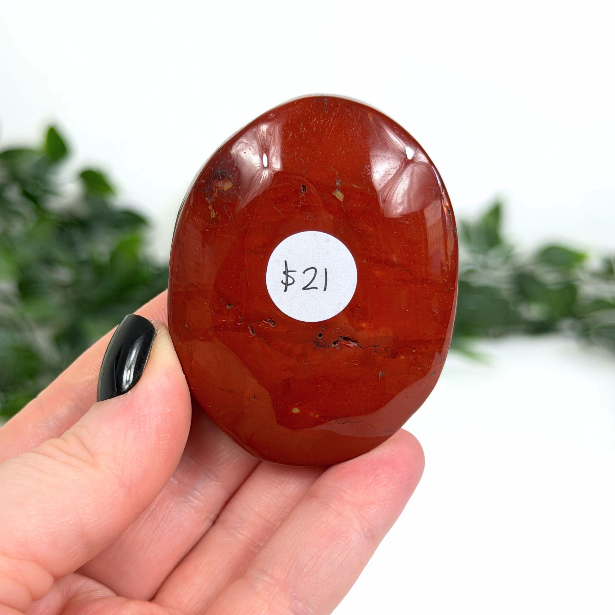 Red mookaite palm stone held in a hand with a price tag label, against a blurred green and white background.