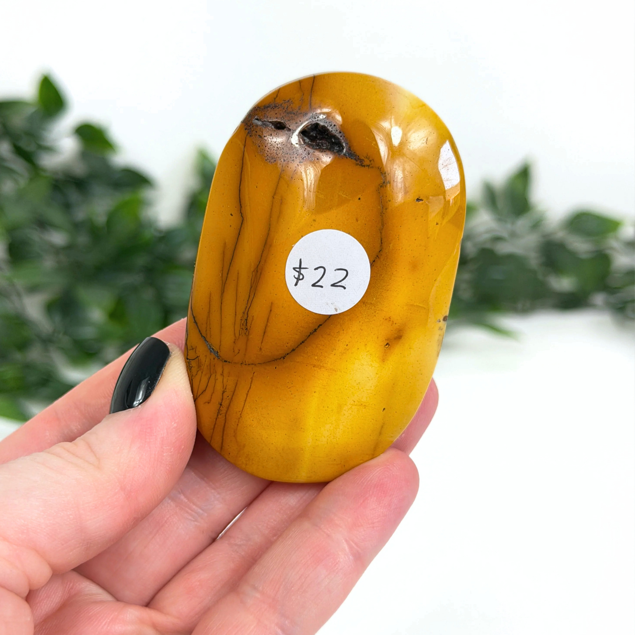 Hand holding a yellow mookaite palm stone with a label on a blurred green background