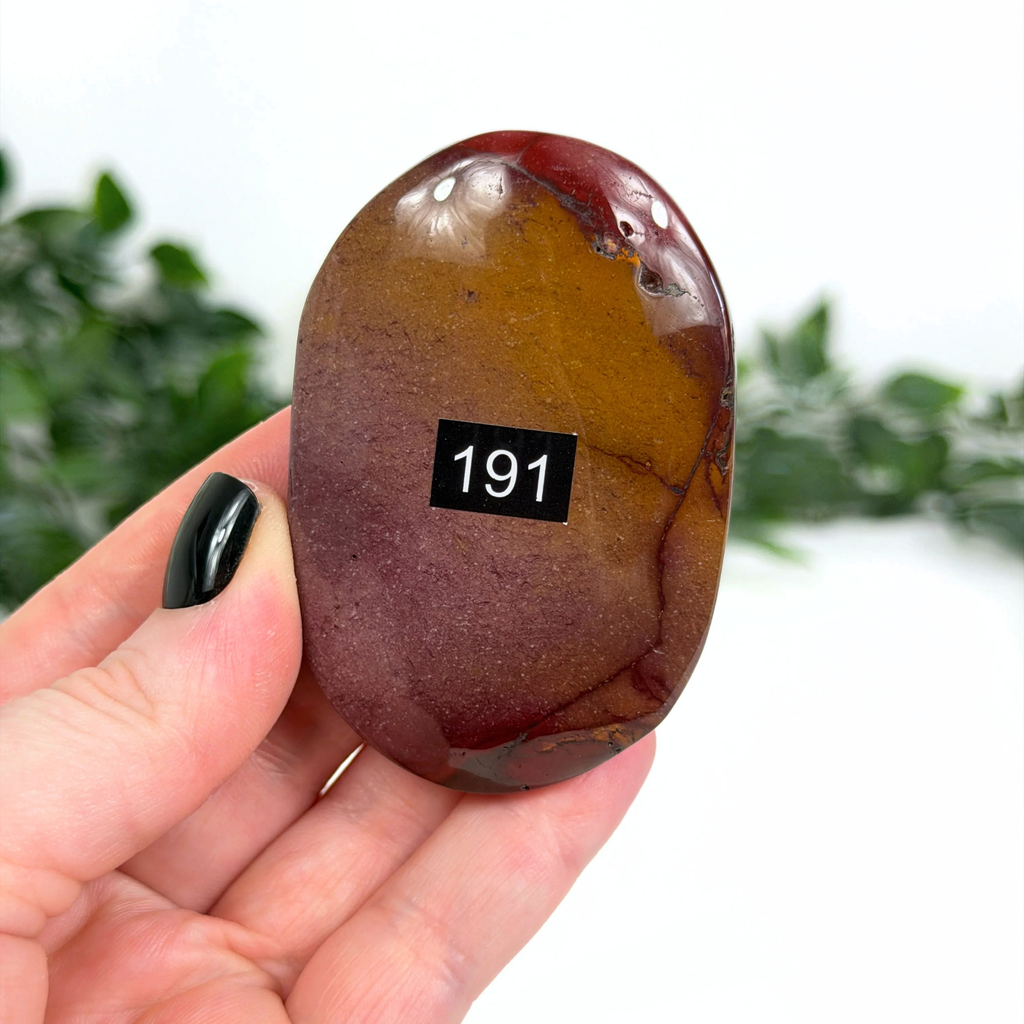 Hand holding a mookaite palm stone with a black label on a blurred green background