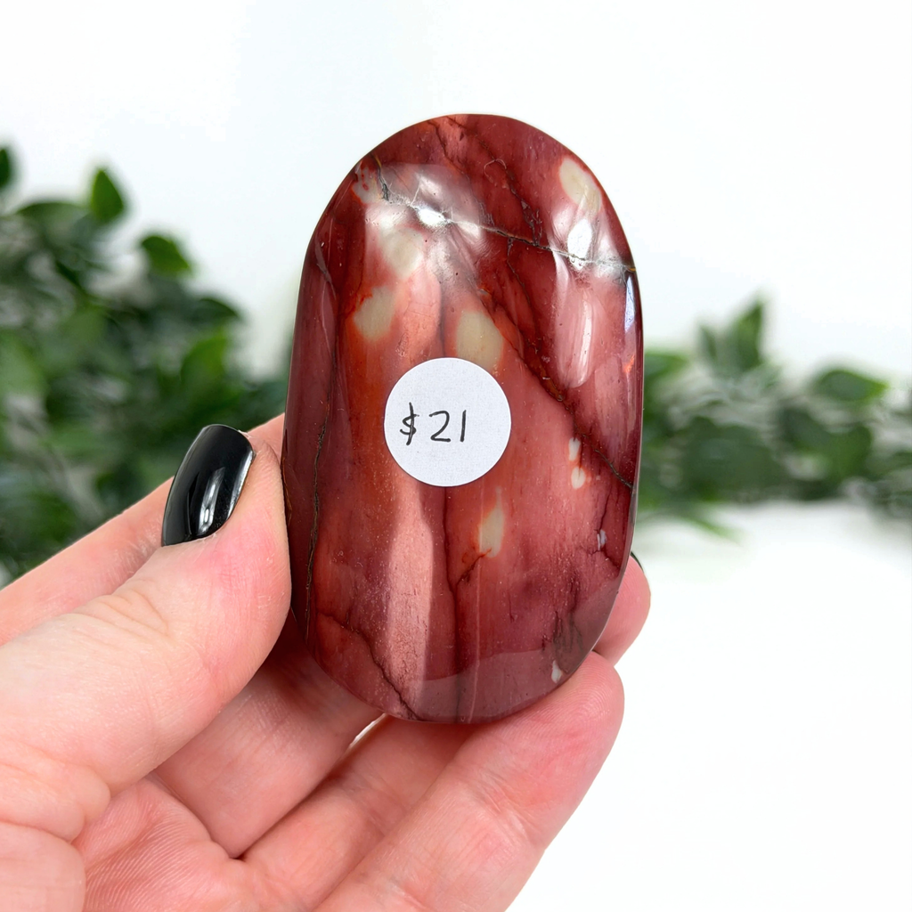Hand holding a red mookaite palm stone with a label on a blurred green background