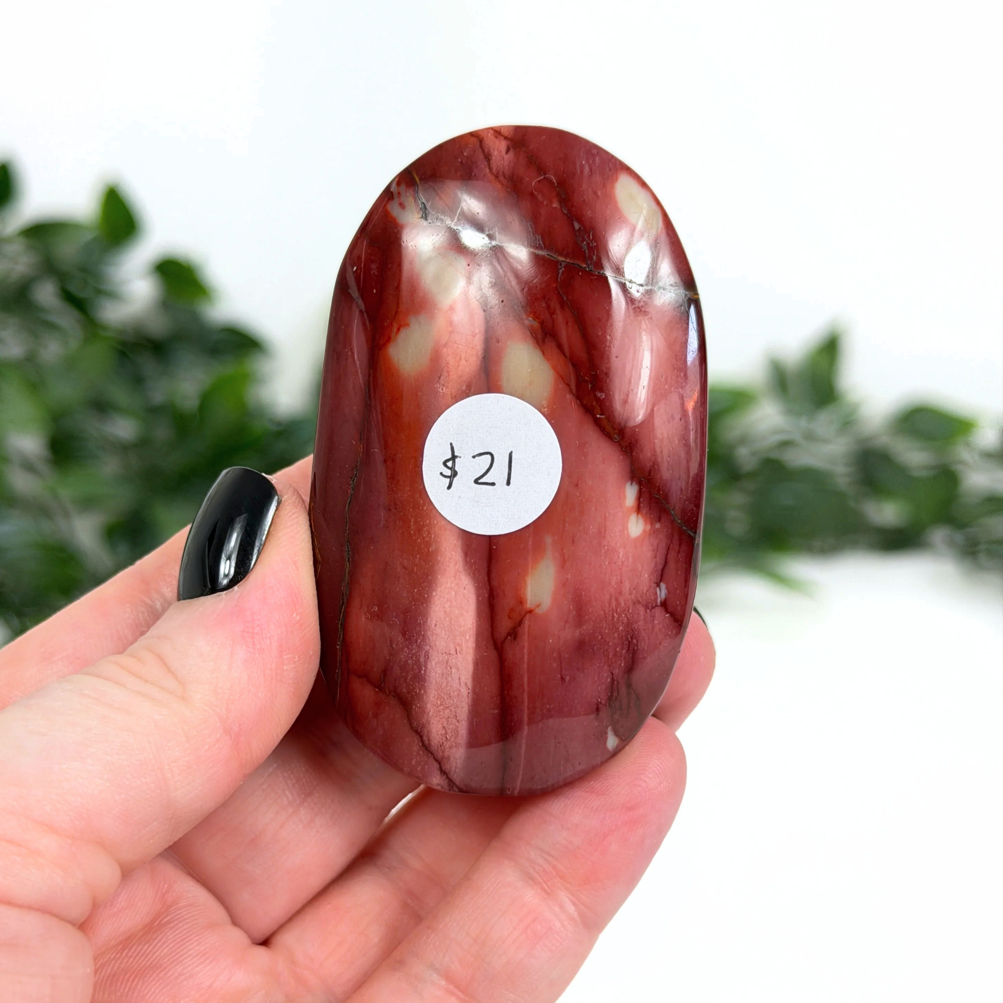 Hand holding a red mookaite palm stone with a label on a blurred green background