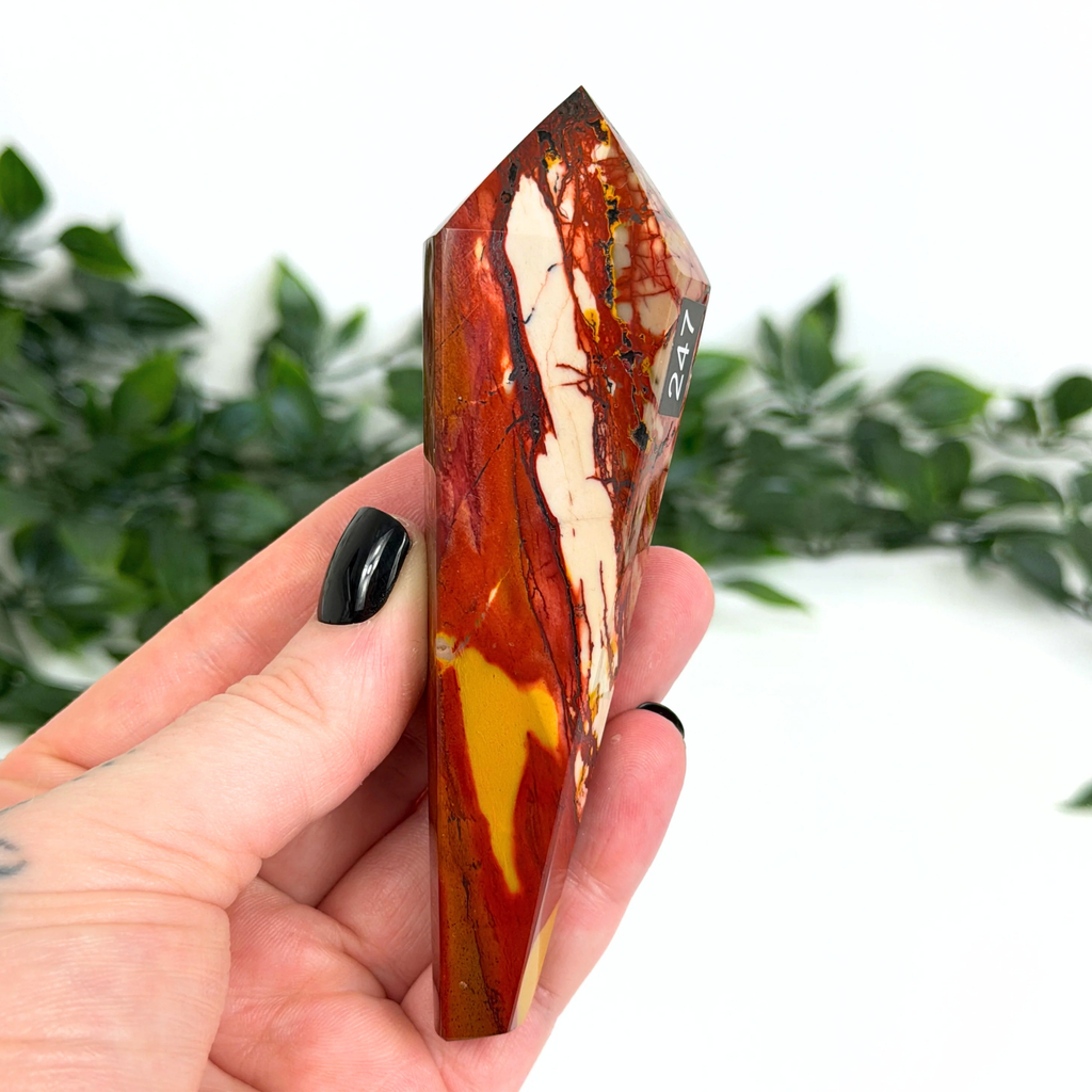 Hand holding a mookaite crystal wand with a blurred green foliage background