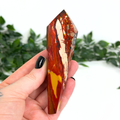 Hand holding a mookaite crystal wand with a blurred green foliage background