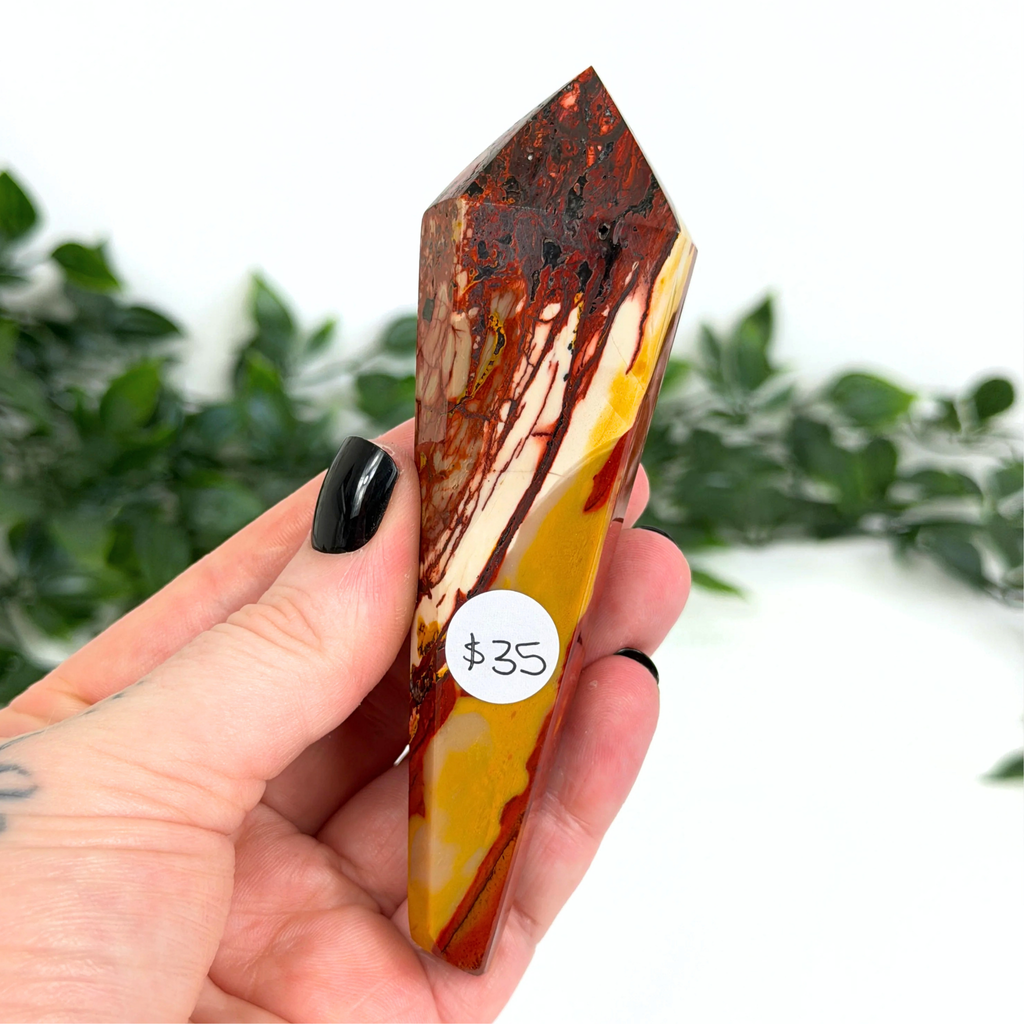 Hand holding a mookaite crystal wand with a $35 label against a blurred green foliage background