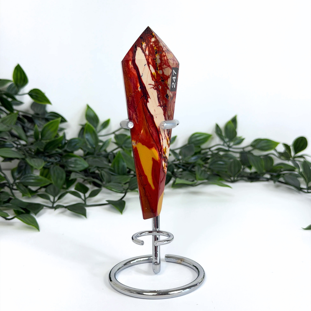 Decorative mookaite crystal wand on a silver stand with green foliage in the background.