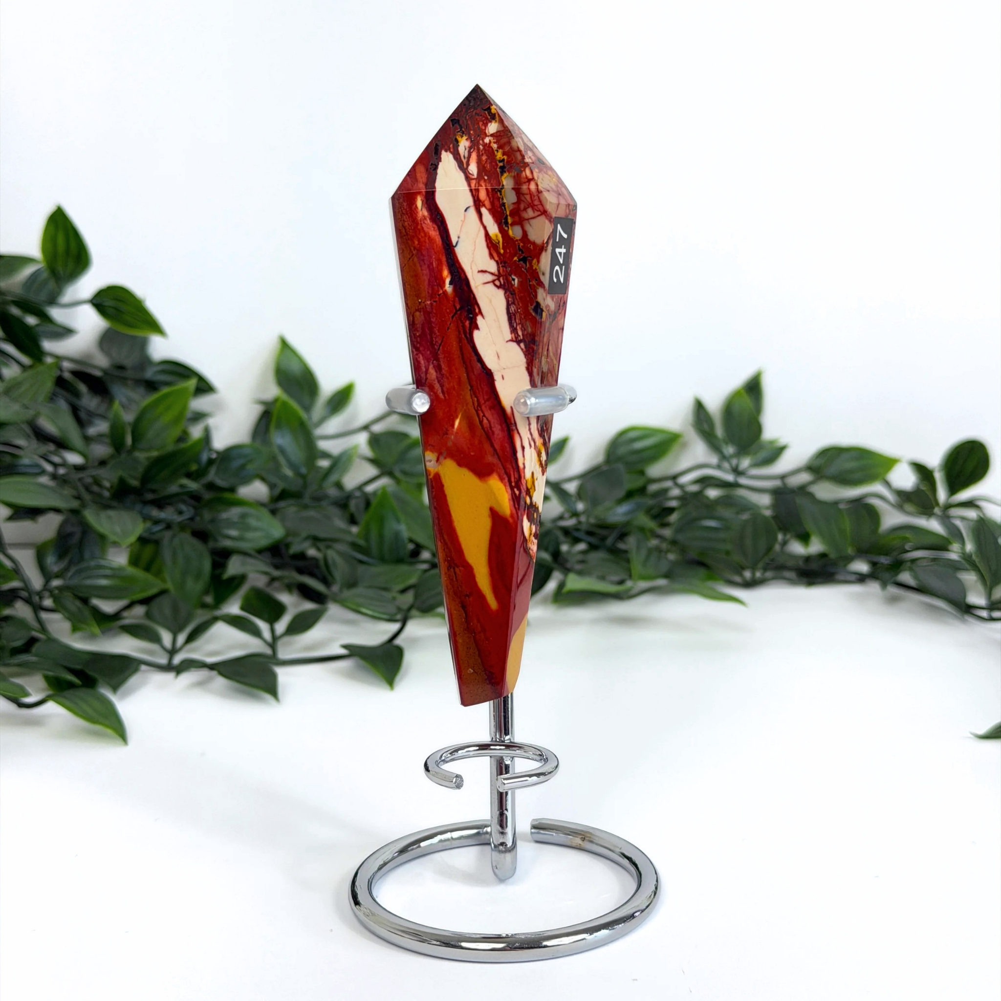 Decorative mookaite crystal wand on a silver stand with green foliage in the background.