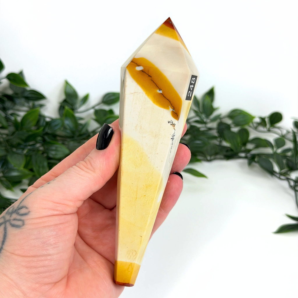 Hand holding a mookaite crystal wand against a white leafy background.