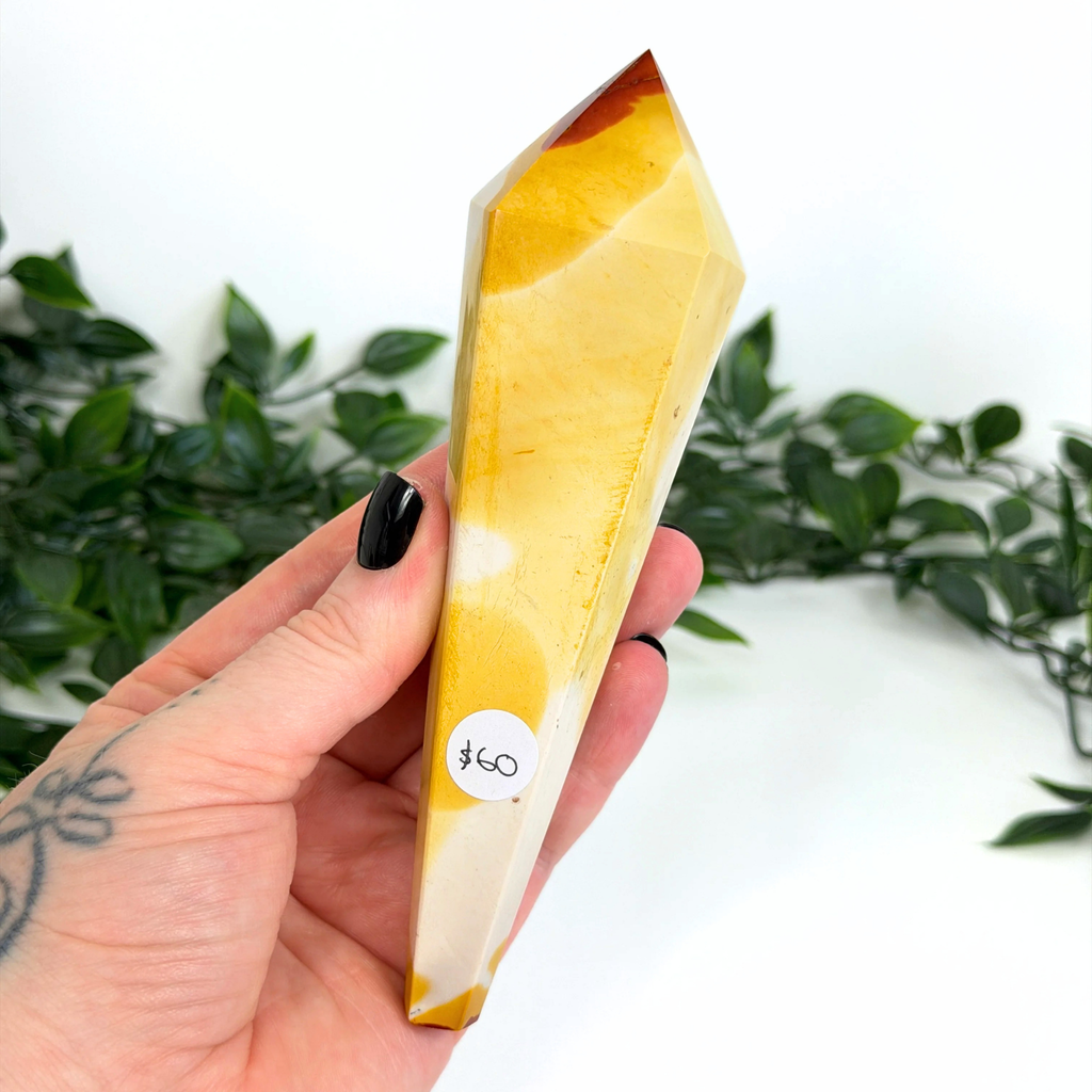 Hand holding a mookaite crystal wand with green foliage in the background