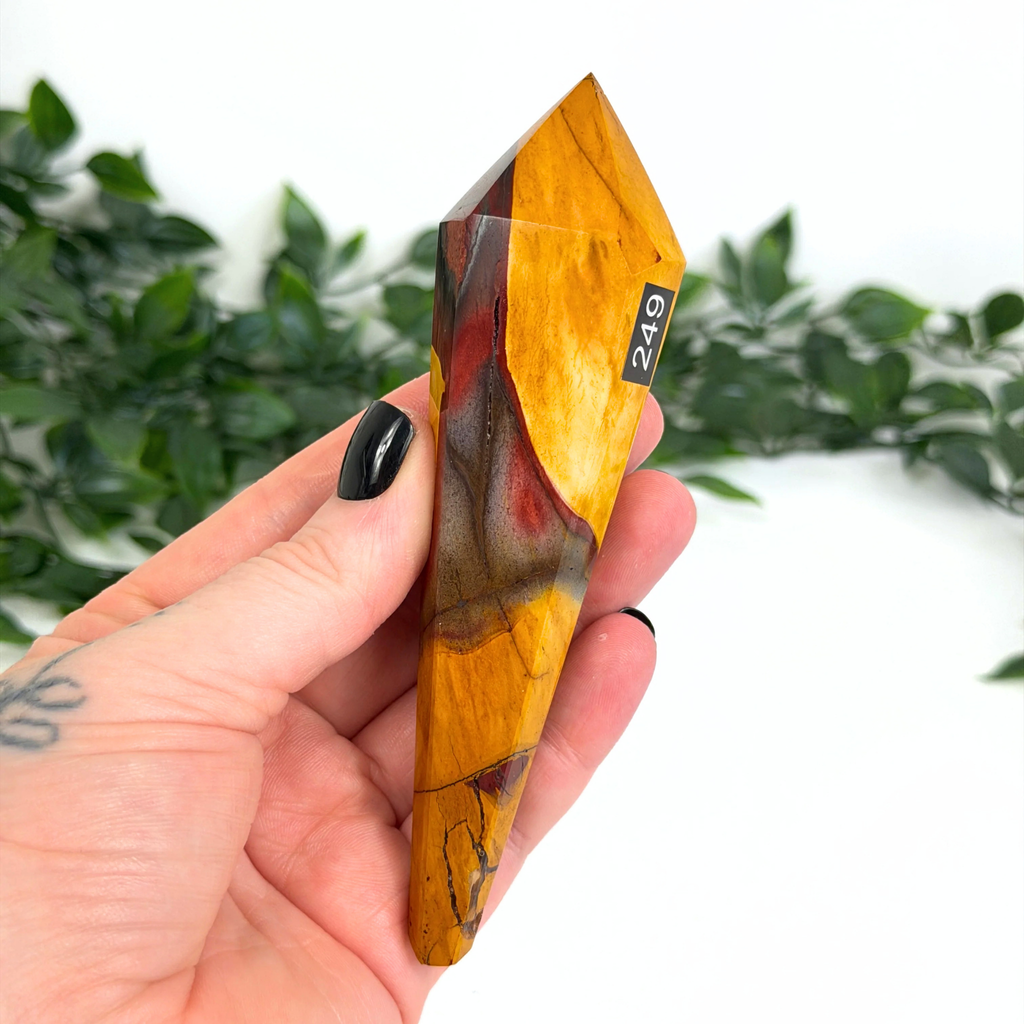Hand holding a mookaite crystal wand with a blurred green leafy background