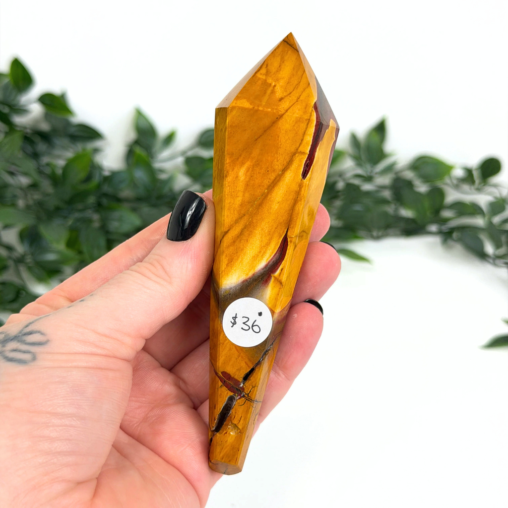 Hand holding a yellow mookaite crystal wand with a blurred green plant background