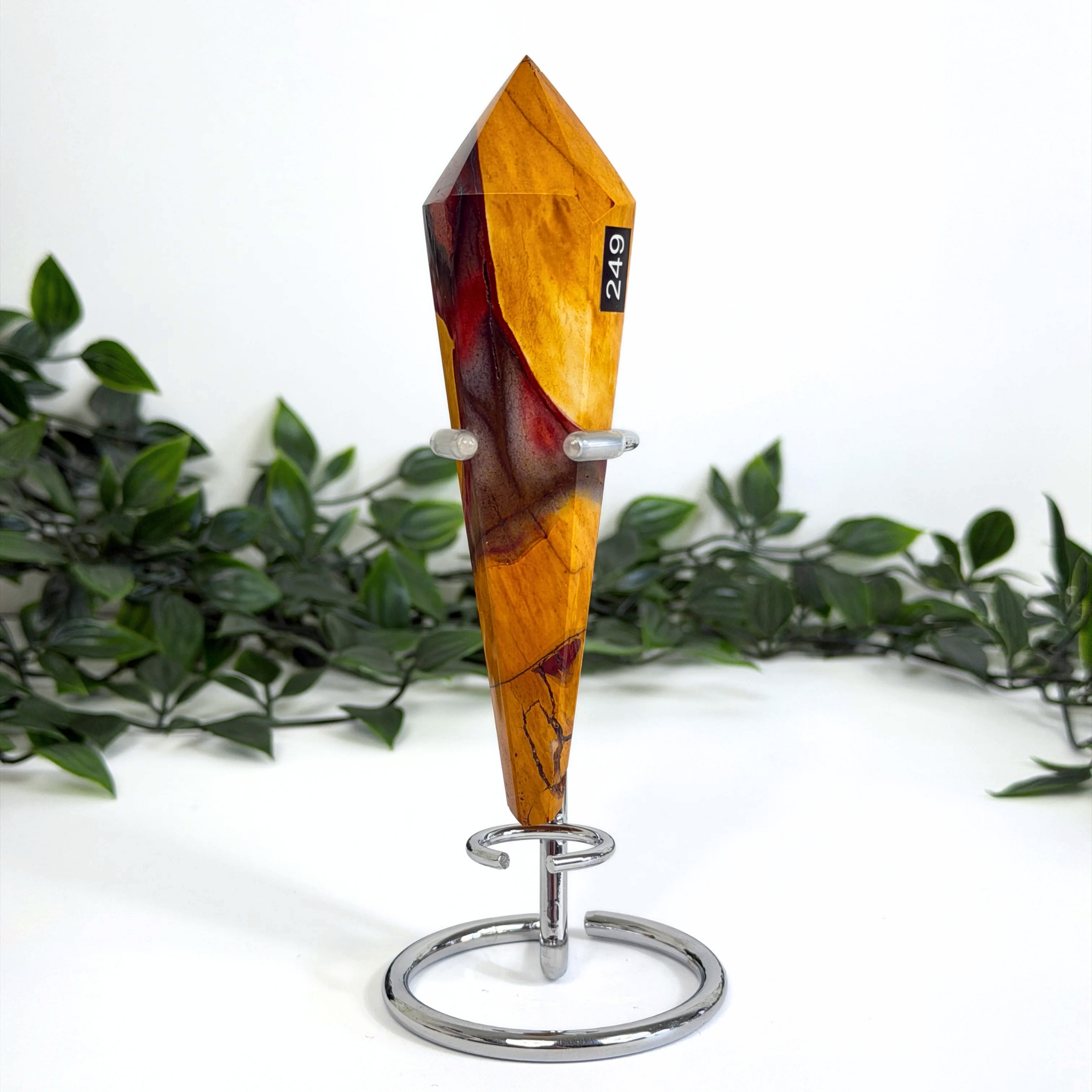 Decorative mookaite crystal wand on a silver stand with a white background and greenery