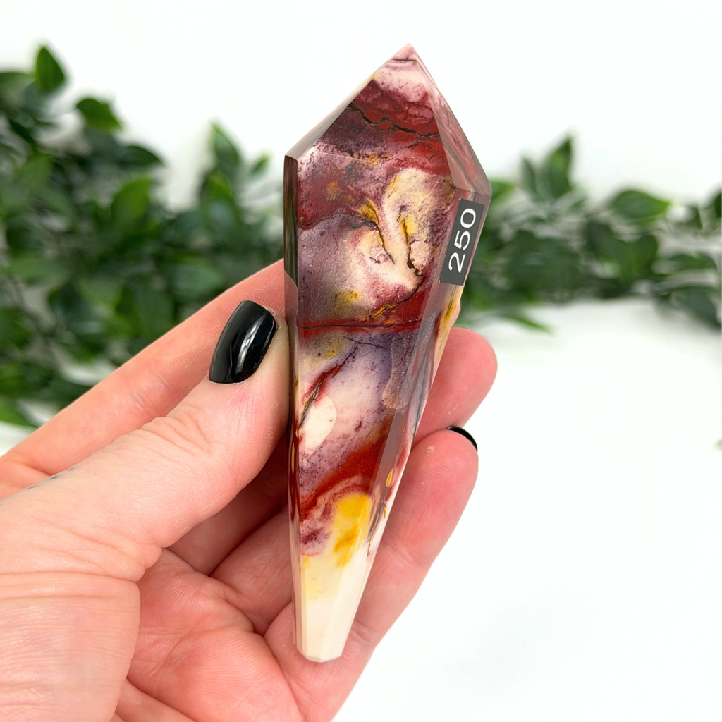 Hand holding a mookaite crystal wand with '250' marking against a blurred green foliage background
