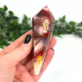 Hand holding a mookaite crystal wand with '250' marking against a blurred green foliage background