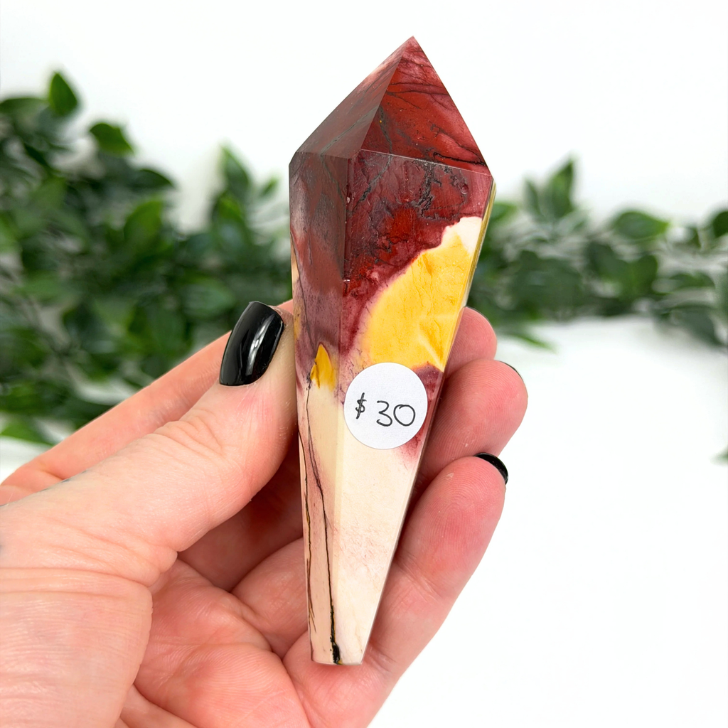 Hand holding a mookaite crystal wand with a price tag labeled $30 against a blurred greenery background