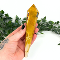 Hand holding a mookaite crystal wand with '251' marking against a white background with green leaves.