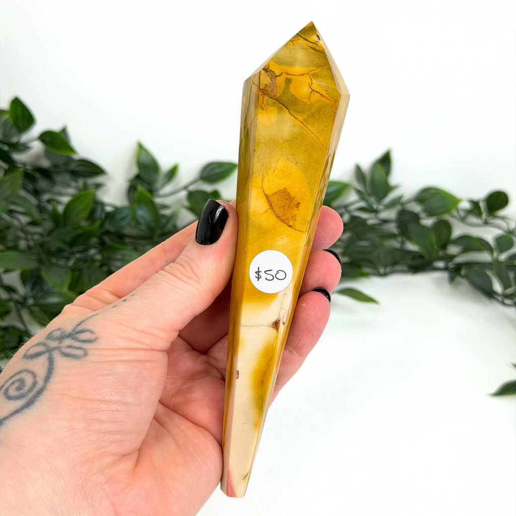 Hand holding a yellow mookaite crystal wand with a '50' label against a green leafy background