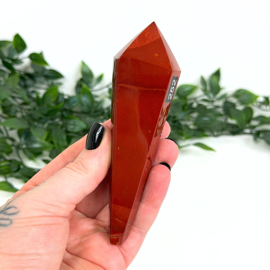 Hand holding a mookaite crystal wand with a blurred green plant background