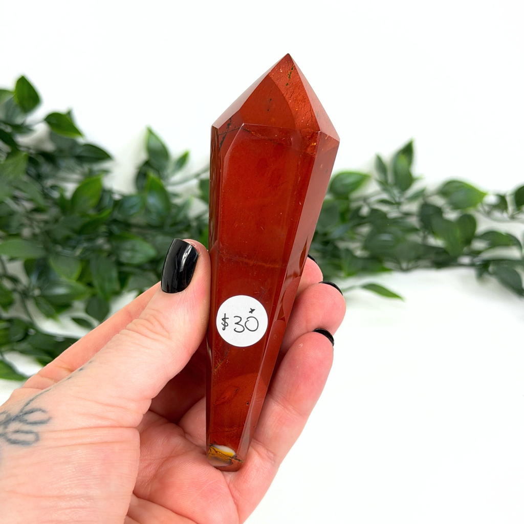 Mookaite crystal wand held by a hand with a blurred green leafy background