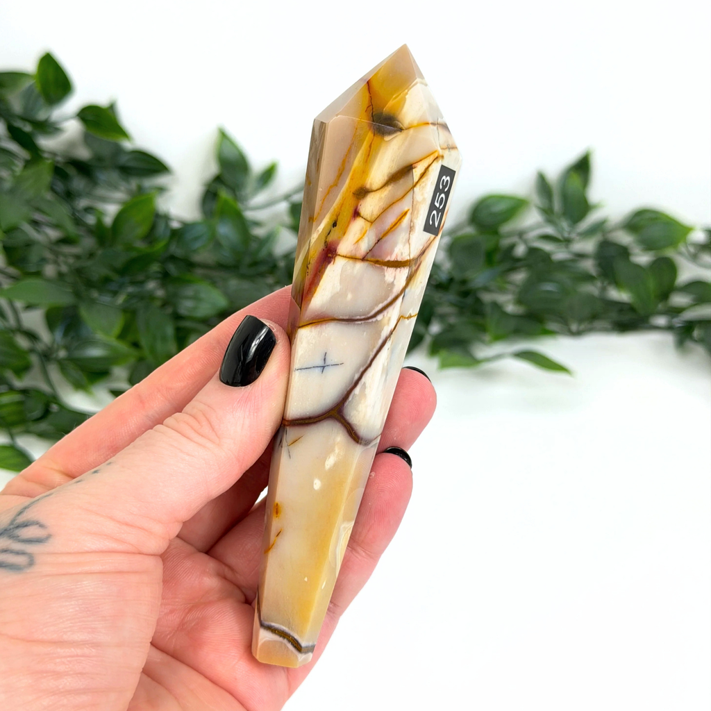 Hand holding a mookaite crystal wand with '253' marking against a blurred green leafy background