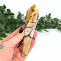 Hand holding a mookaite crystal wand with '253' marking against a blurred green leafy background