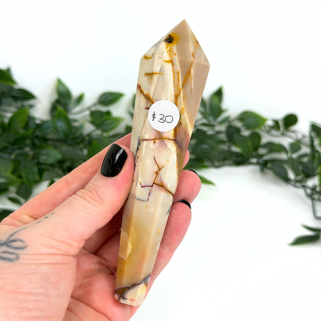 Hand holding a mookaite crystal wand with a white background and green foliage