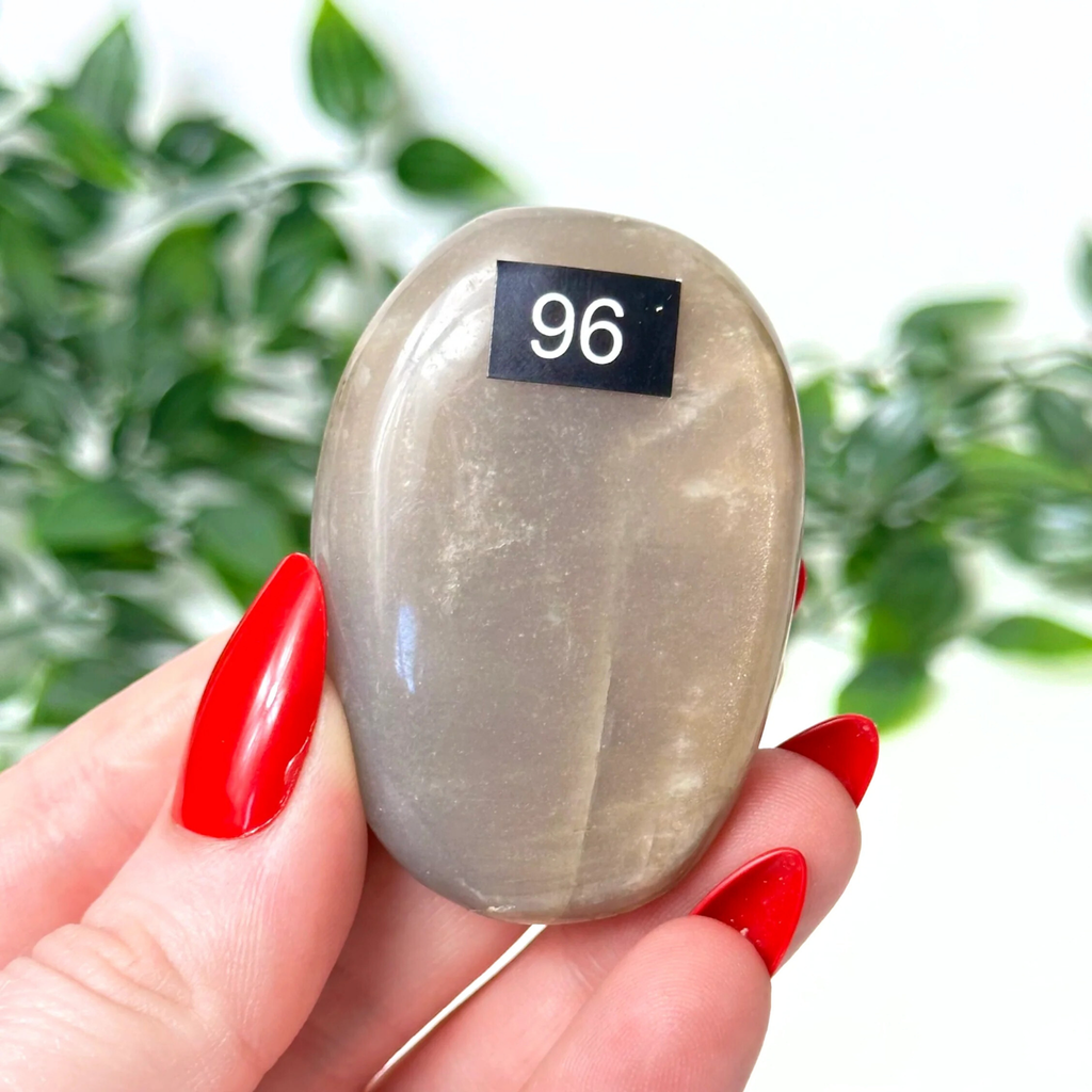 Hand holding a moonstone palm stone against a blurred green leafy background