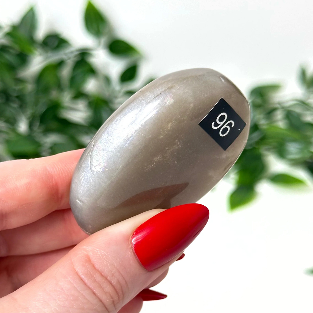 Hand holding a moonstone palm stone against a blurred green leafy background