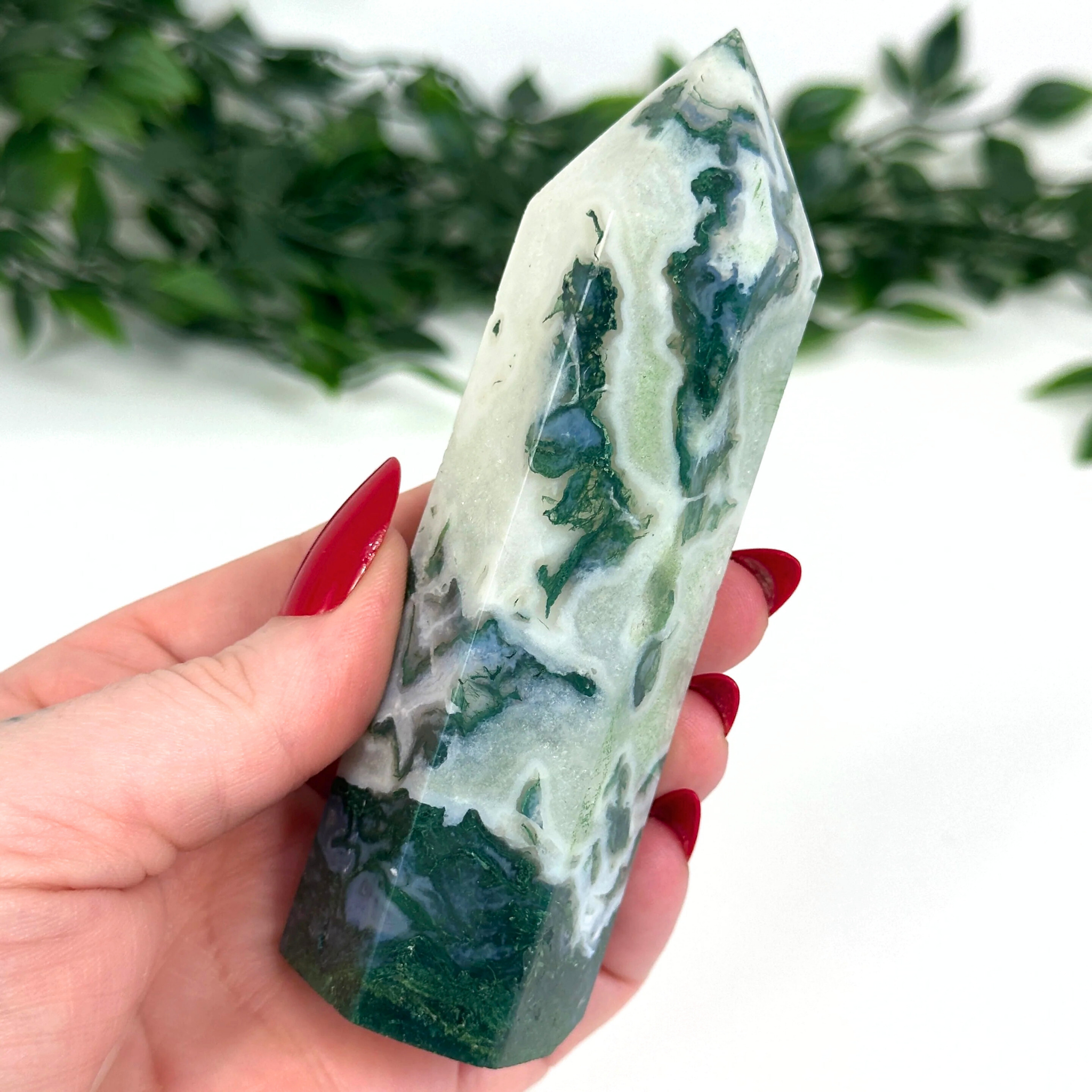 Hand holding a green and white moss agate crystal tower with a blurred green leafy background