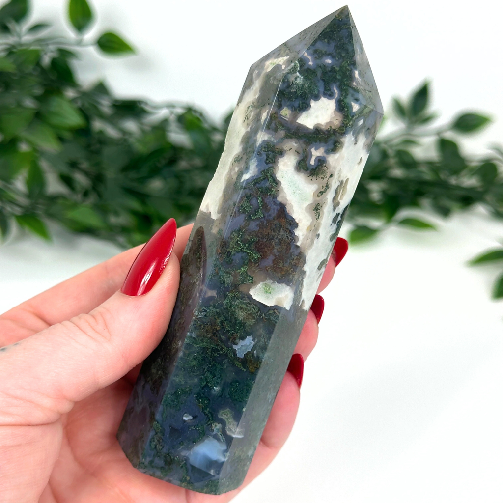 Hand holding a moss agate crystal tower with a green and white pattern against a blurred natural background