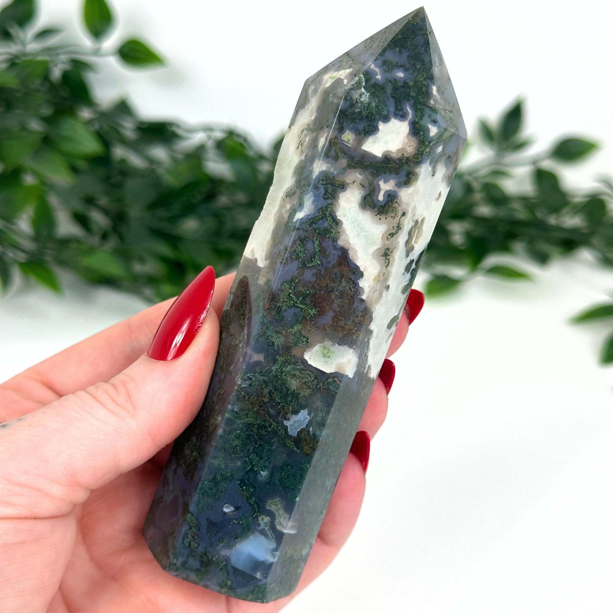 Hand holding a moss agate crystal tower with a green and white pattern against a blurred natural background