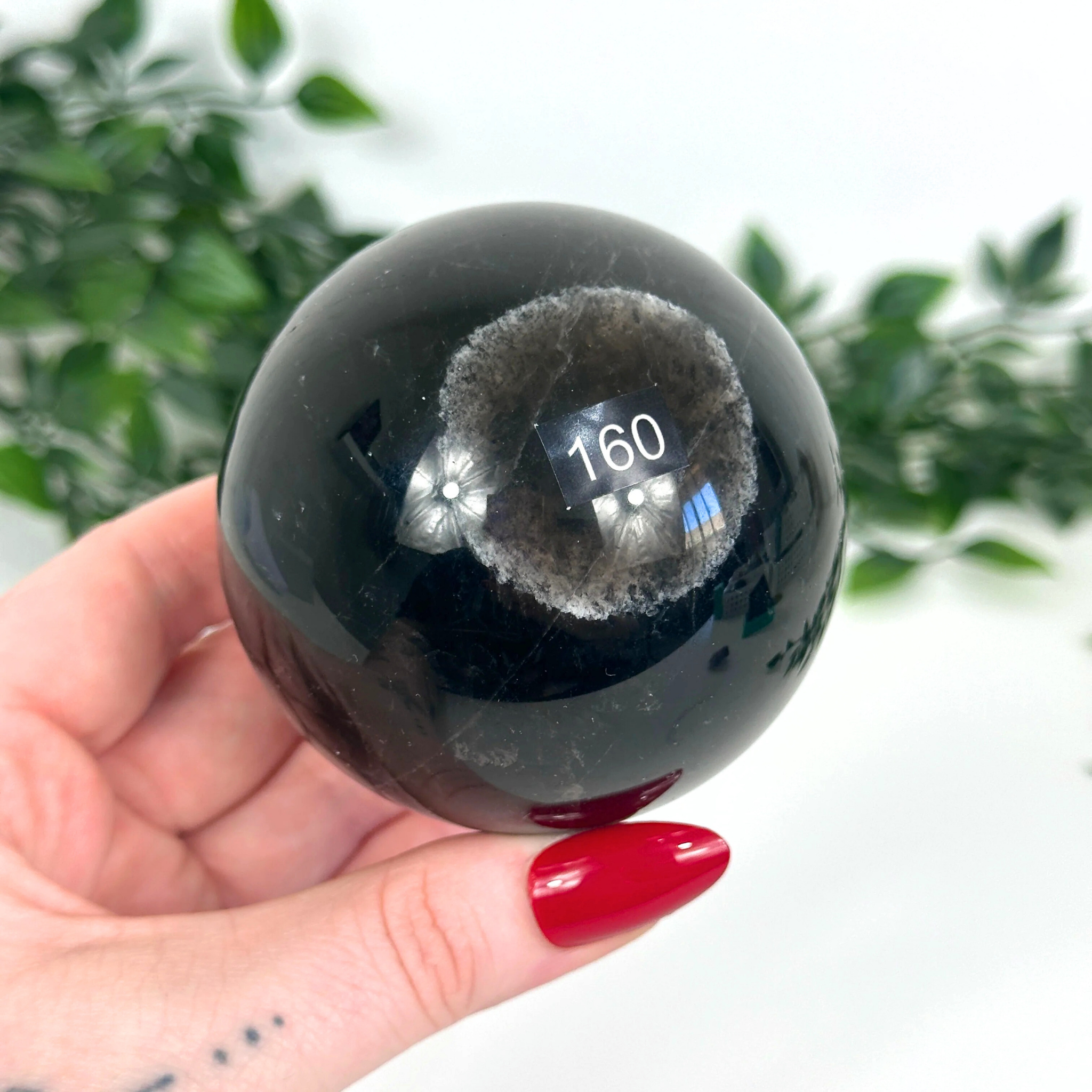 Smoky quartz crystal ball held in a hand with red nail polish, against a blurred green leafy background.