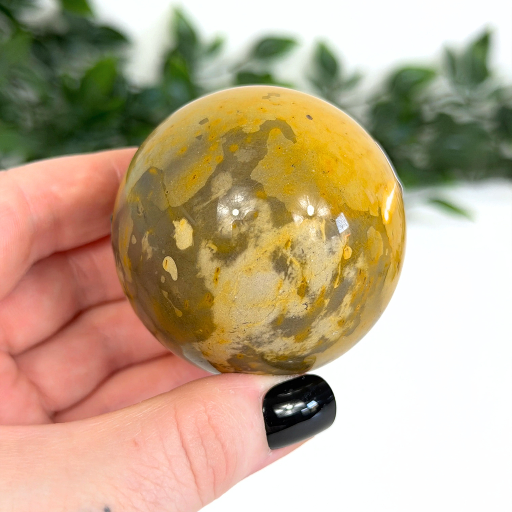 Hand holding a yellow and brown mookaite sphere with a blurred green background