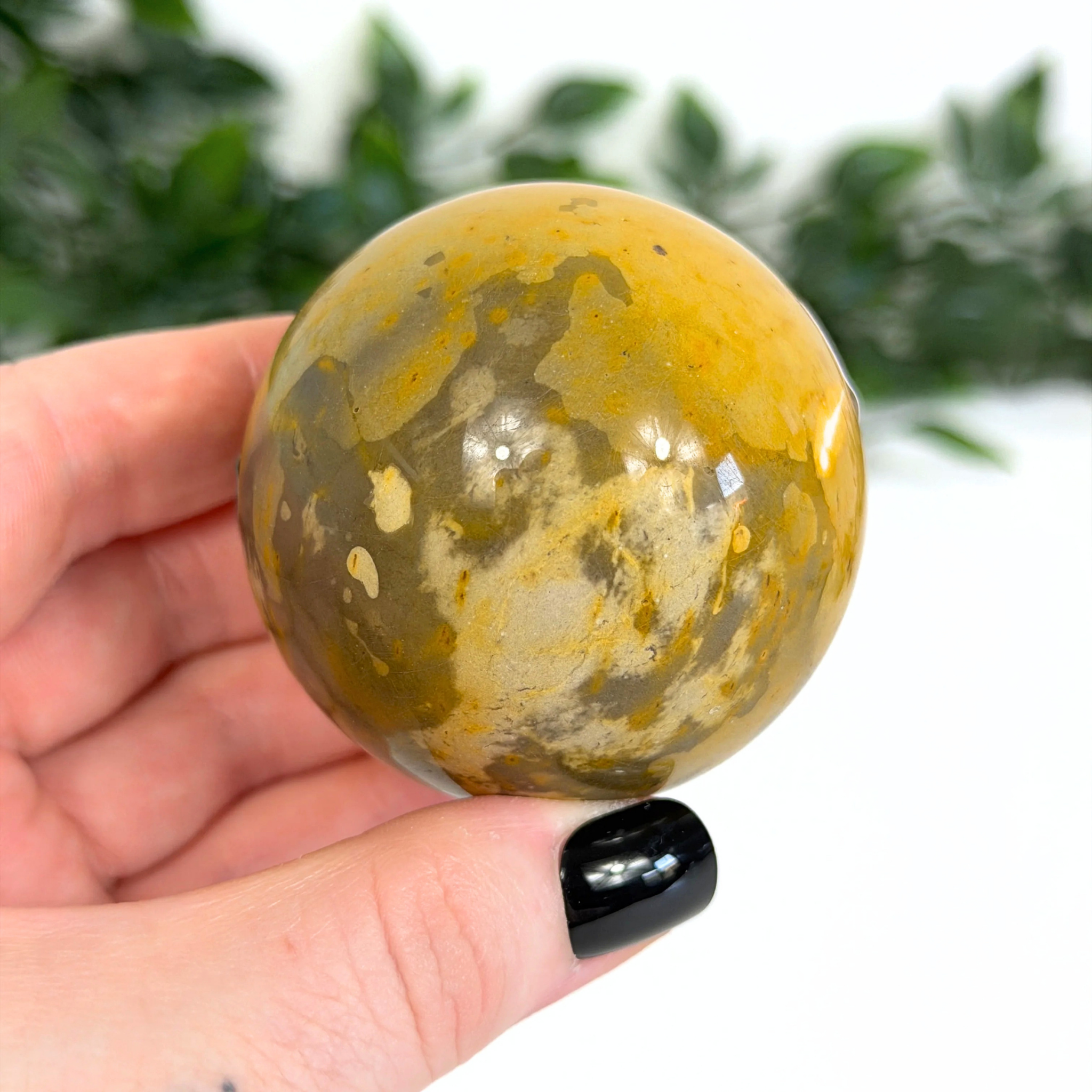 Hand holding a yellow and brown mookaite sphere with a blurred green background