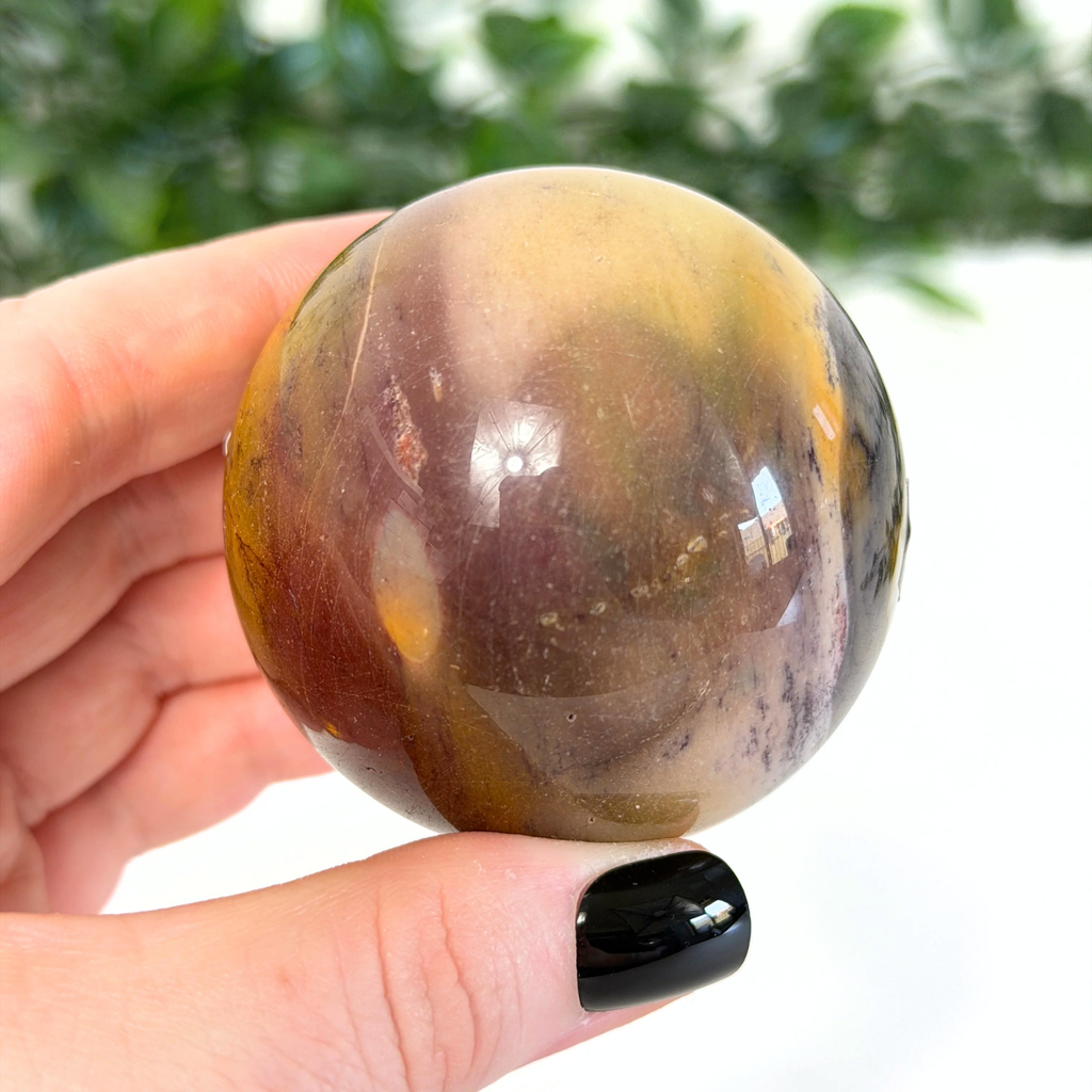 Hand holding a mookaite crystal sphere with a blurred green background