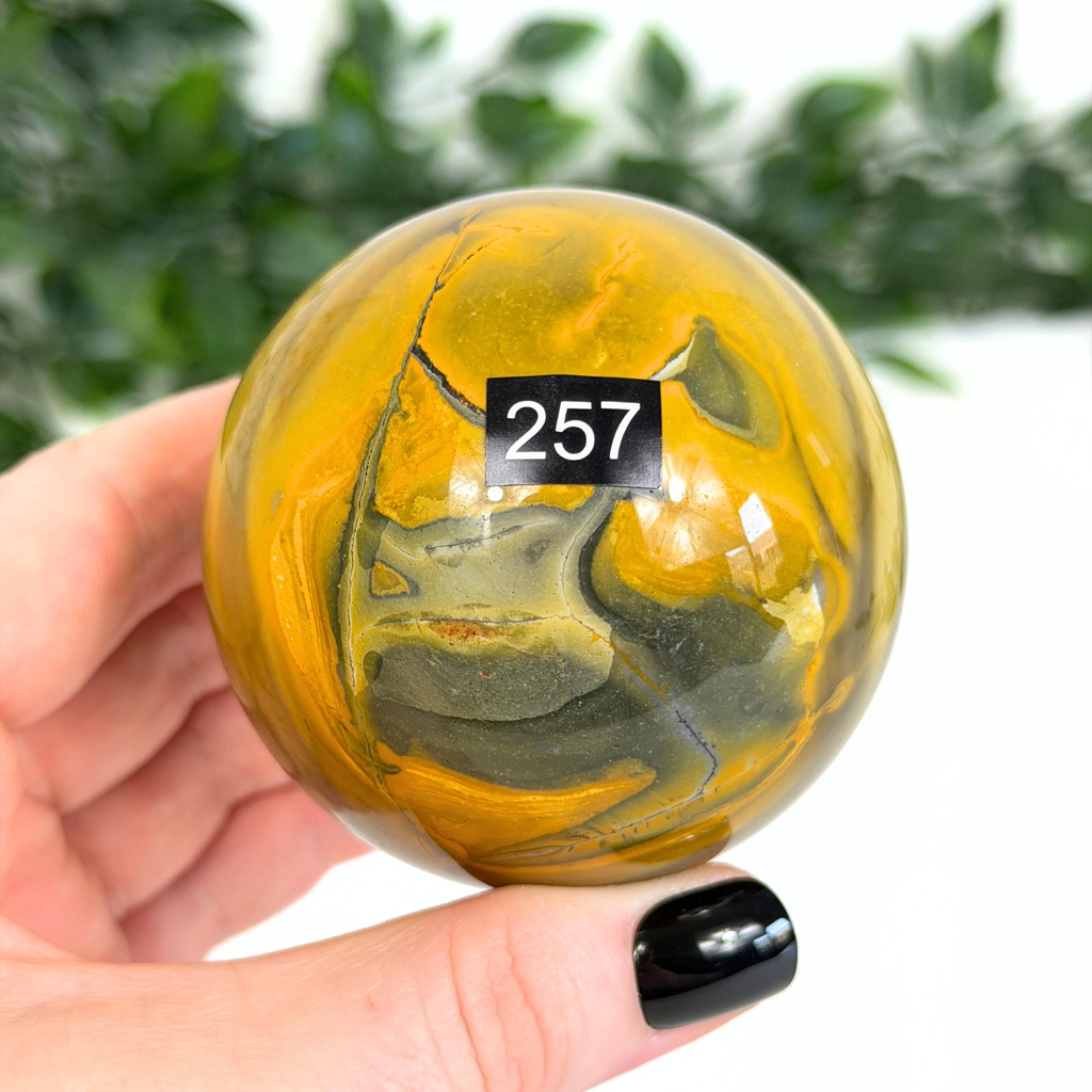 Marbled yellow and gray mookaite sphere held in a hand with black nail polish, against a blurred green background.