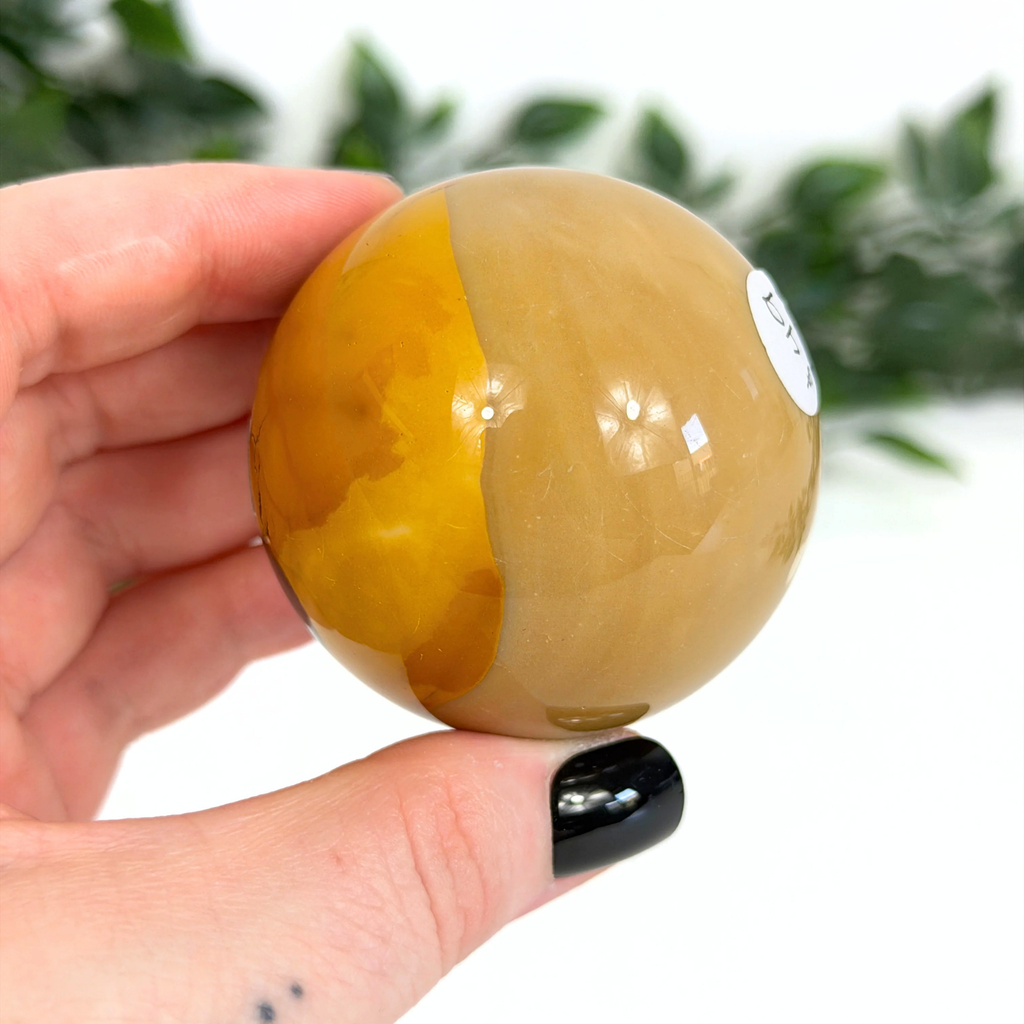 Hand holding a brown and yellow mookaite sphere with a blurred green background