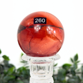 Red mookaite sphere with a number 260 label on a clear stand against a blurred green background
