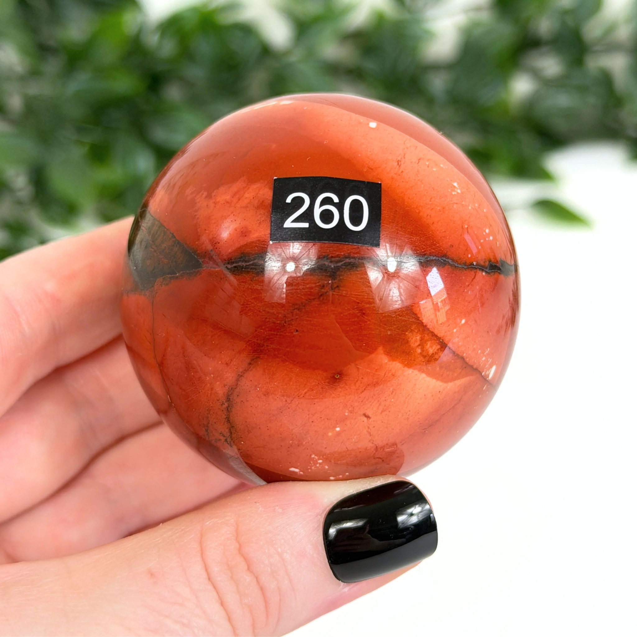 Red nookaite crystal sphere with a black label on a hand against a blurred green background