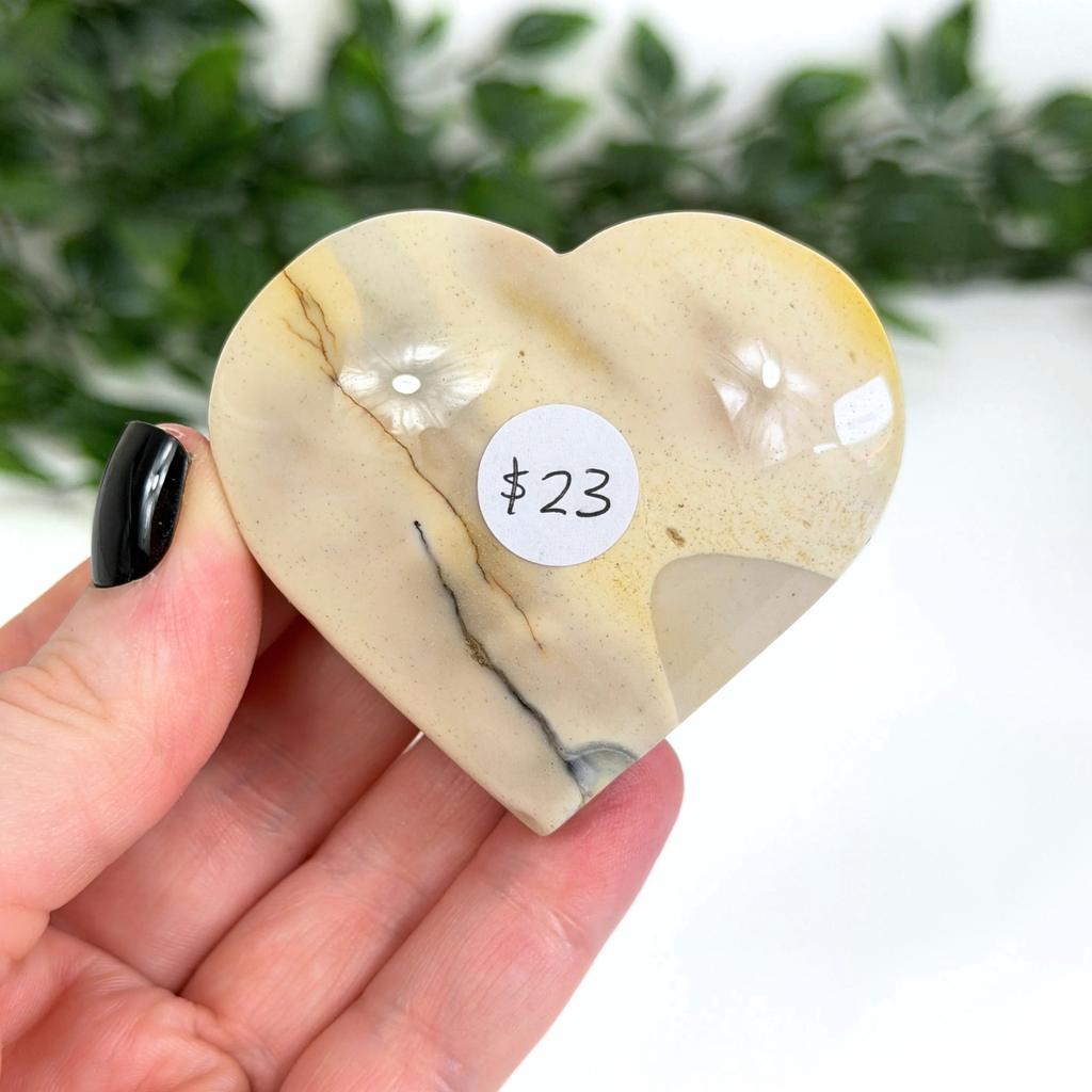 Heart-shaped mookaite stone held in a hand with a price tag label, against a blurred greenery background.