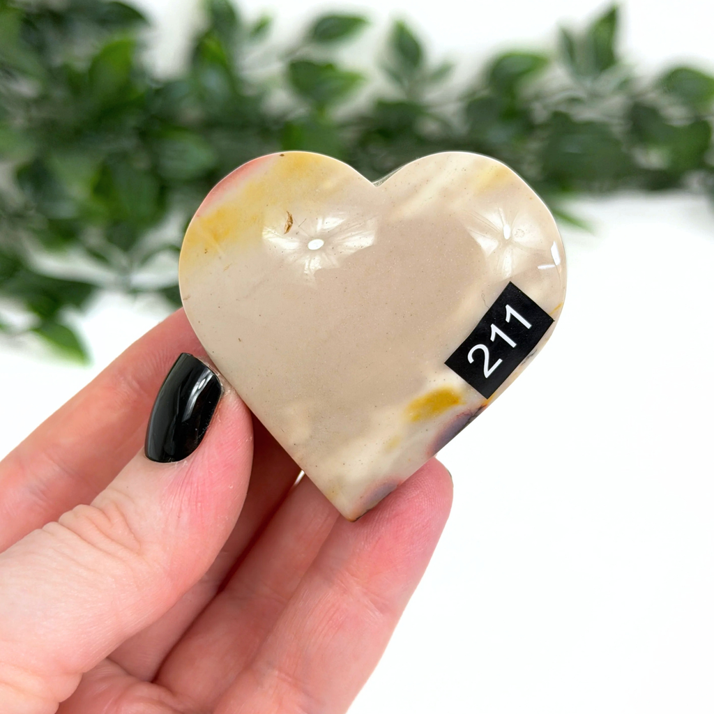 Heart-shaped mookaite stone held in a hand with a blurred green leafy background