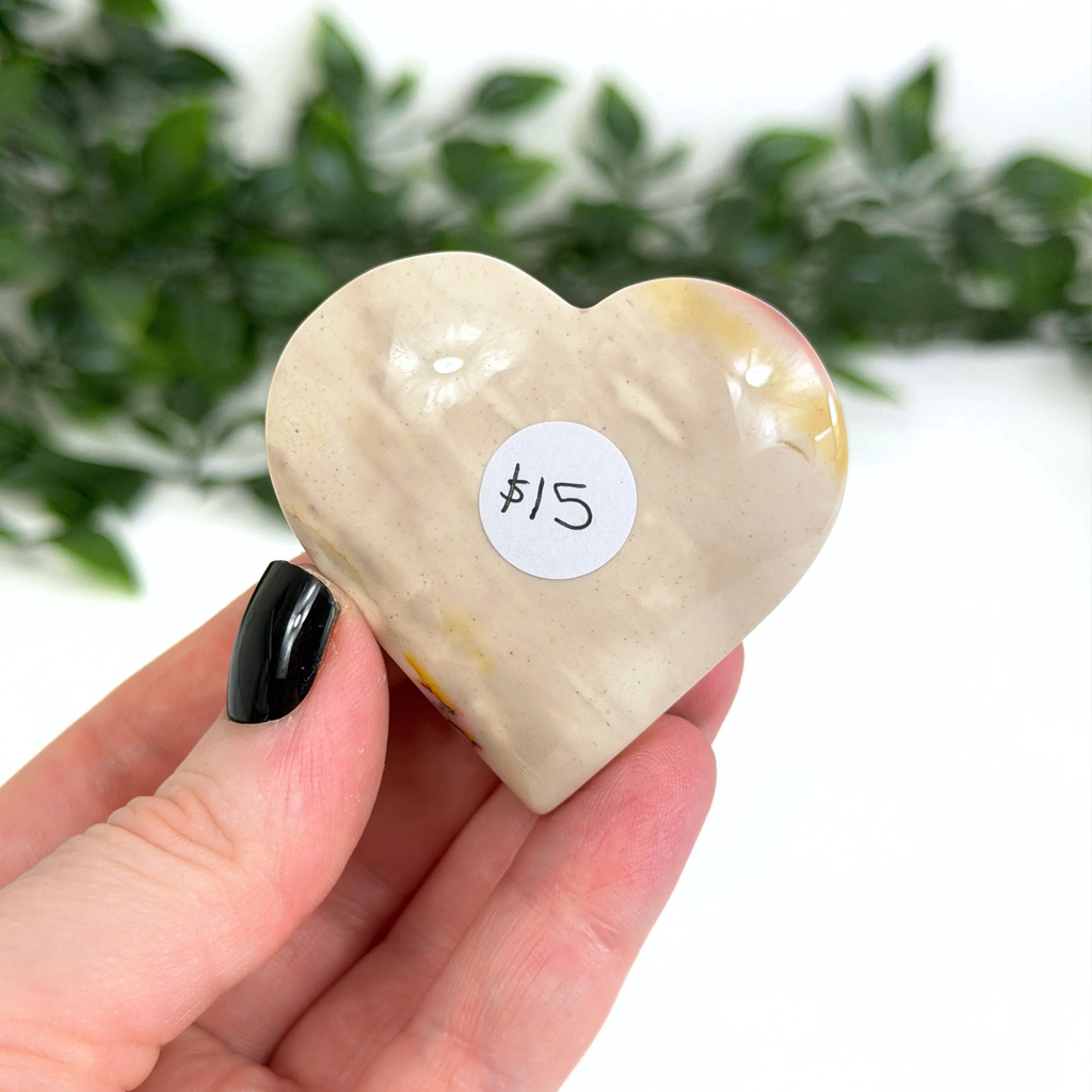Heart-shaped mookaite crystal held in a hand with a price tag label, against a blurred green leafy background.