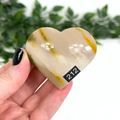 Heart-shaped mookaite stone held in a hand with a blurred green leafy background