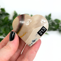 Heart-shaped mookaite crystal with '213' marking held in a hand against a blurred green background