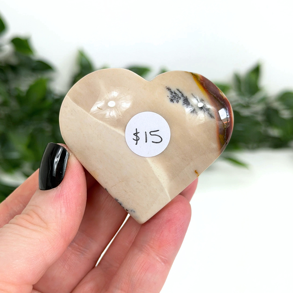 Heart-shaped mookaite crystal with a price tag held by a hand against a blurred green background