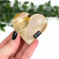 Heart-shaped mookaite crystal held in a hand with a blurred green leafy background
