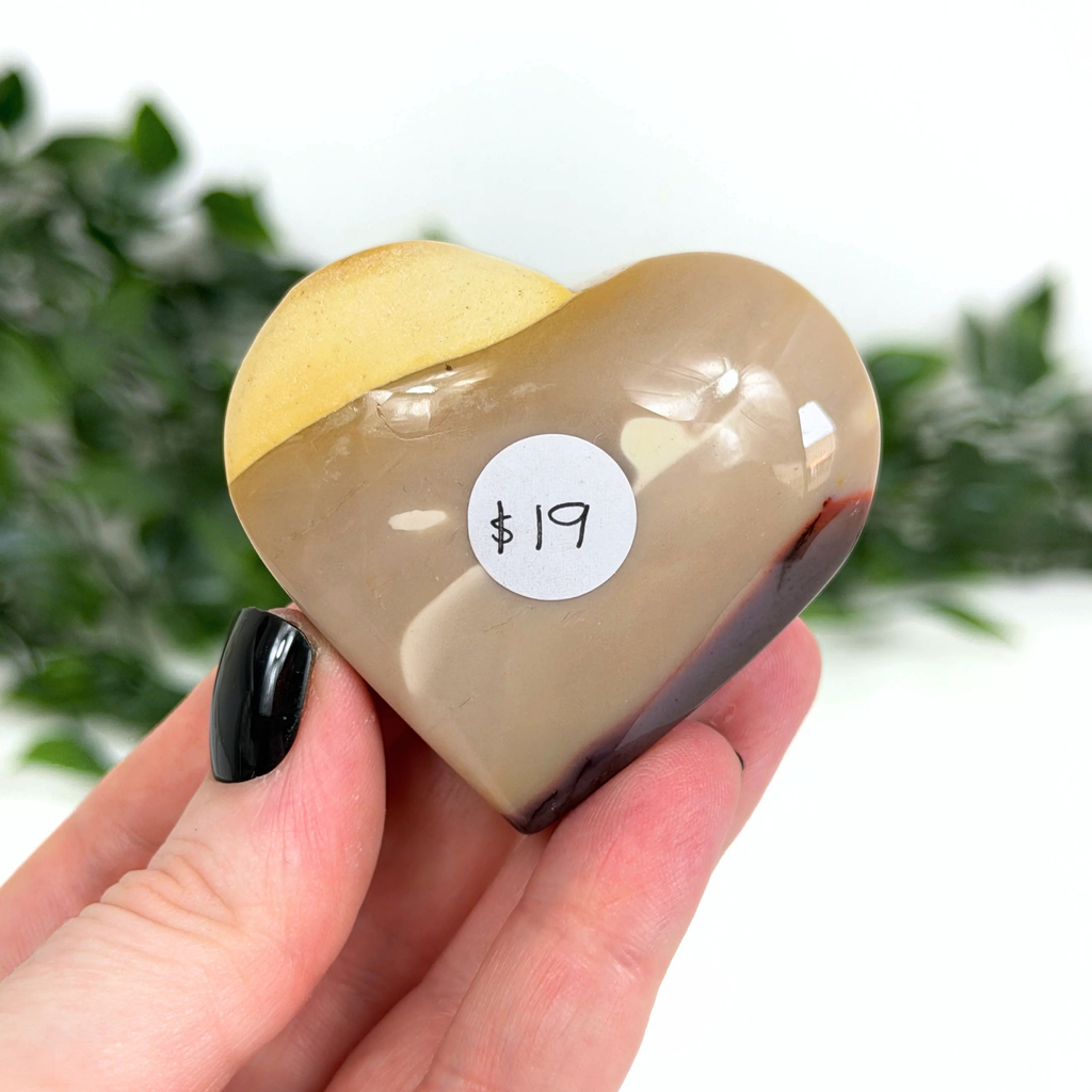 Heart-shaped mookaite crystal with a price tag held in a hand against a blurred green background